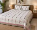 A bed dressed in a cream quilted coverlet with a pink and green floral trellis pattern, matching pillowcases, and a decorative pink and green border, styled in a warm, modern bedroom setting.